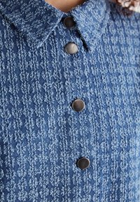 Blue patterned denim shirt featuring a textured fabric, a classic collar, and black buttons along the front.