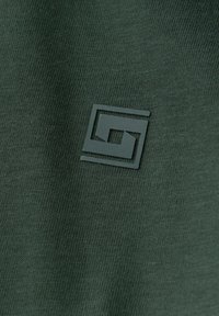 Zielona dzianina ma teksturowaną powierzchnię z wyraźnym szarym logo. Logo składa się z geometrycznych, stylizowanych kształtów.