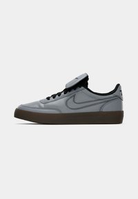 Grå Nike-sko med svart swoosh-logo, svarte lisser og mørkebrune såler, vist i sidevinkel på hvit bakgrunn.