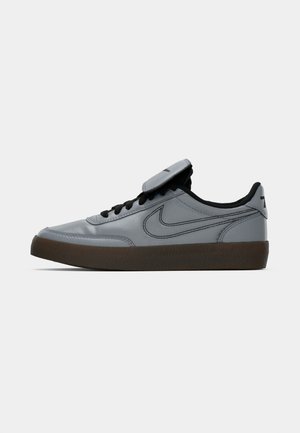 Zapatilla Nike gris con logotipo Swoosh negro, cordones negros y suela marrón oscuro, mostrada en vista lateral sobre un fondo blanco.