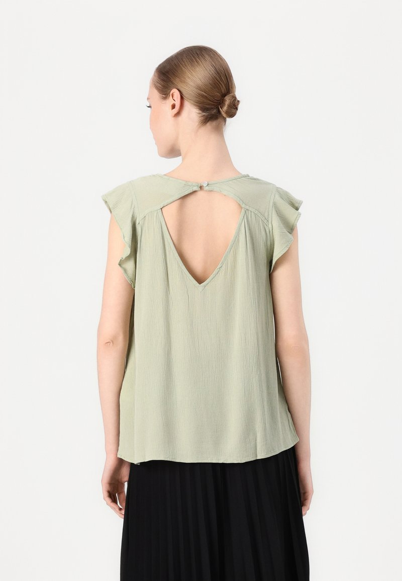 Lys grøn kortermet bluse med V-rygg og volanger. Stoffet har en teksturert finish, med knappelukking bak.