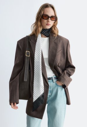 Jonge vrouw draagt een oversized bruine blazer, wit topje, lichtblauwe spijkerbroek, polkadot sjaal, amberkleurige zonnebril en draagt een donkere schoudertas.