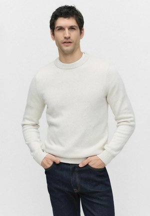 Pull en maille beige clair avec un col rond, des poignets et un ourlet côtelés, associé à un jean bleu foncé. Texture lisse et coupe standard.