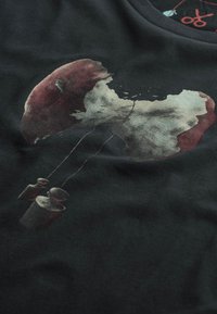 Donkere stof met een gedrukt ontwerp van een rode en witte paddenstoel die uit elkaar splitst, opgehangen aan twee kleine gewichten. Gladde textuur.