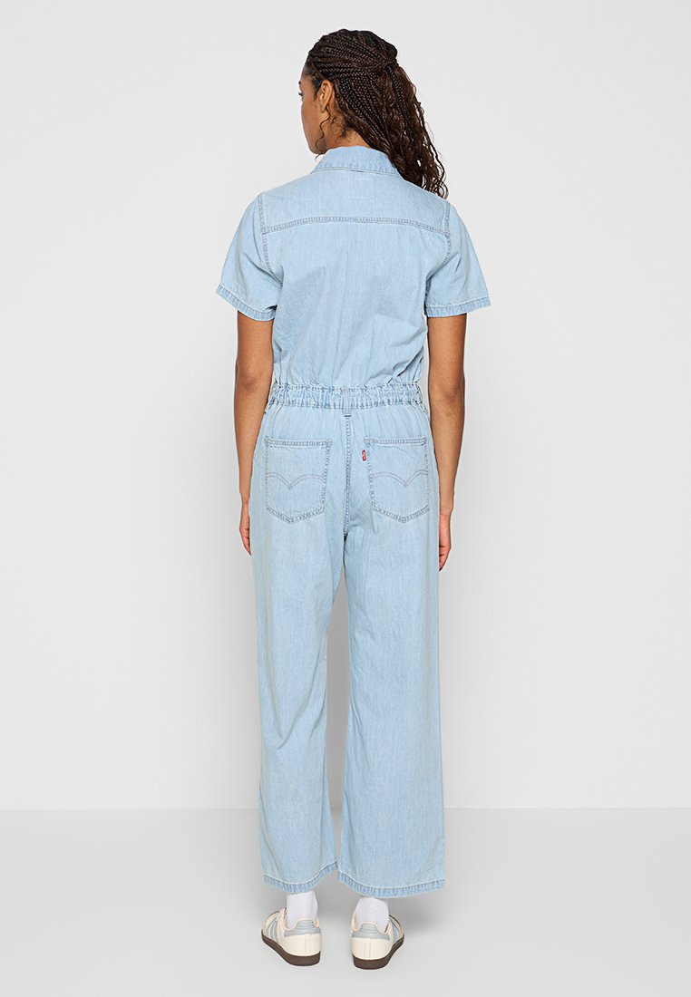 Combinaison en denim bleu clair avec un col, des manches courtes, une taille élastique et des jambes larges. Dispose de poches arrière et de coutures discrètes.