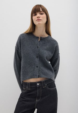 Grijze gebreide cardigan met ronde hals, volledige mouwen en voorlopende knoopsluiting; gecombineerd met hoge taille donkere spijkerbroek.