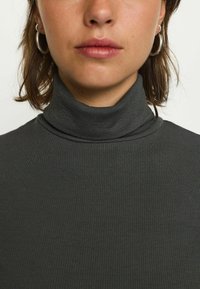 Dunkelgrauer gerippter Rollkragenpullover mit naheliegendem Kragen. Verfügt über eine glatte Textur und ein minimalistisches Design. Sichtbare silberne Creolen.