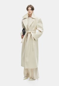 Beige overjas met grote kraag, ceintuur in de taille en wijde mouwen, gedragen over een bijpassende outfit. Houdt een kleine zwarte clutch vast. Glad textuur.