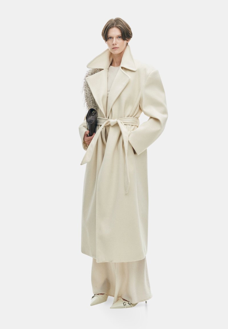 Beige overjas met grote kraag, ceintuur in de taille en wijde mouwen, gedragen over een bijpassende outfit. Houdt een kleine zwarte clutch vast. Glad textuur.