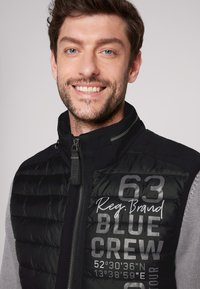 Gilet imbottito nero con chiusura a zip davanti, maniche grigie e grafiche stampate che includono "Reg. Brand", "63" e coordinate. Materiale testurizzato.