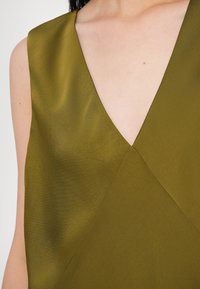 Olívazöld, ujjatlan felső V-nyakkivágással, sima textúrával és ferdén futó varrással. Nincsenek látható minták vagy fém részletek.