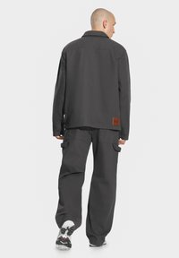 Szary płaszcz wykonany z wytrzymałego materiału z klasycznym kołnierzykiem. W połączeniu z luźnymi szarymi spodniami cargo, które mają boczne kieszenie i brązową metkę marki.