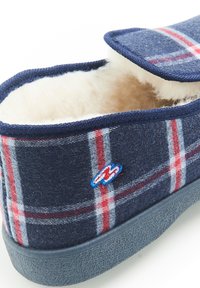 Chausson à carreaux bleu marine avec intérieur en laine, présentant des rayures rouges et blanches, une semelle grise texturée et un logo brodé bleu sur le côté.