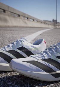 Paar weiße und schwarze Laufschuhe auf einer gepflasterten Außenfläche mit weißer Linie unter klarem blauem Himmel.