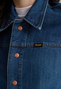 Džinsinis švarkas su klasikiniu apykakle, vario spalvos sagomis ir juodu etiketės logotipu. Ryški mėlyna spalva su matomomis siūlėmis.