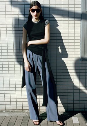 Femme portant des lunettes de soleil foncées, un haut noir, une longue écharpe noire, un pantalon gris à jambes larges et des sandales, debout contre un mur carrelé au soleil.