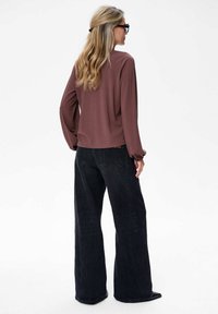 Vrouw met lang blond haar, draagt schildpadschaalbril, een mauve langemouwen top en wijde zwarte jeans, zittend met de rug naar de camera op een witte achtergrond.