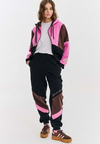 Młoda kobieta ubrana w różowo-brązową i czarną bluzę kolorową i dopasowane dresy oraz brązowe sneakersy na białym tle.