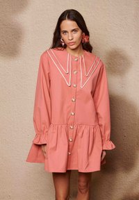 Ροζ βαμβακερό φόρεμα με μεγάλο γιακά, λευκή επένδυση και κουμπιά στο μπροστινό μέρος. Διαθέτει στενές στρώσεις και φουσκωτά μανίκια με βολάν στις μανσέτες.