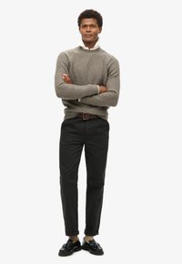Pull en tricot gris clair avec texture côtelée, pantalon noir tapered et chaussures habillées noires. Le mannequin se tient les bras croisés sur un fond blanc.