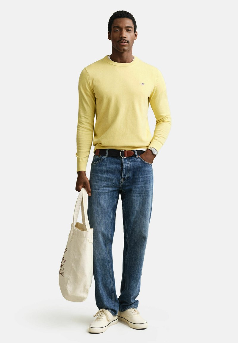 Mann trägt gelben Pullover, blaue Jeans, weiße Sneakers, hält eine beige Tragetasche, steht vor weißem Hintergrund.