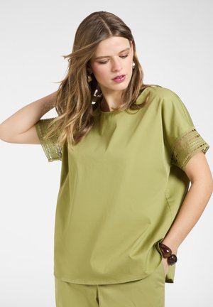 Woman with light brown hair wearing a loose olive green short-sleeve top with lace trim and matching pants, posing with one hand in pocket.