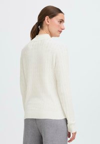 White knit sweater with a textured diamond pattern, ribbed cuffs, and hem. Worn with gray pants, shown from the back.