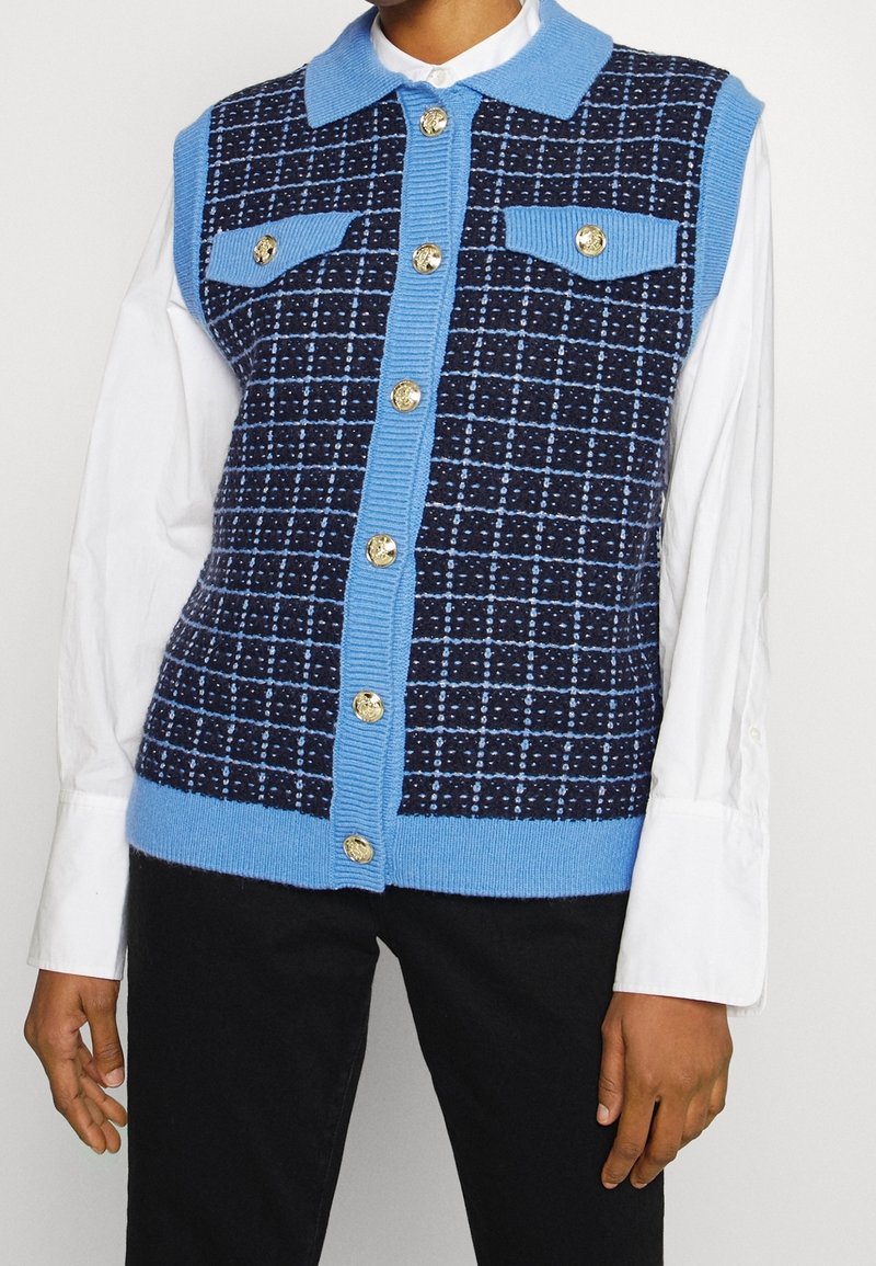 Veste tricotée sans manches à carreaux bleu et bleu marine avec des boutons dorés, portée sur une chemise blanche à manches longues et associée à un pantalon noir.