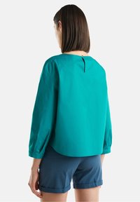 Blusa verde acqua ampia con maniche lunghe, caratterizzata da un dettaglio a chiave sul retro. Abbinata a pantaloni corti blu arrotolati. Texture del tessuto liscia.