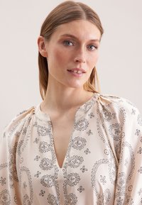 Cremige Bluse mit tiefem Ausschnitt, puffigen Ärmeln und schwarzen Blumenmustern. Hergestellt aus einem glatten, leichten Stoff.