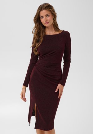 Femme aux longs cheveux ondulés portant une robe midi ajustée à manches longues couleur bordeaux foncé avec une fente latérale, des boucles d'oreilles en or, un collier et des bracelets.