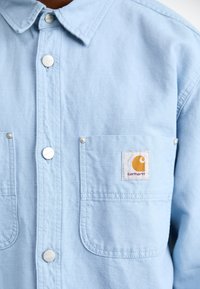 Jasnoniebieska koszula Carhartt zapinana na metalowe zatrzaski, z przednią kieszonką i pomarańczową łatką z logo marki.