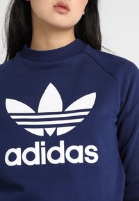 Sweatshirt bleu marine en coton, avec un logo Adidas blanc comprenant trois bandes et un design trèfle sur le devant. Style simple avec col rond.