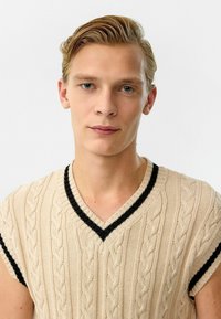 Beige gebreide trui met korte mouwen en V-hals, voorzien van een kabelpatroon en zwarte afwerking langs de halslijn en mouwen.