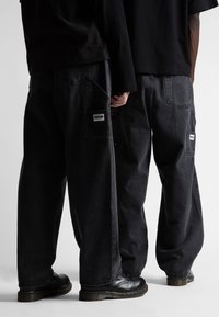 Juodos džinsinės baggio kelnės su didelėmis kišenėmis, logotipo aplikacijomis gale ir šiek tiek nusidėvėjusia tekstūra. Vilkima su juodais batais.