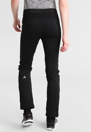 Person trägt schwarze Sporthosen und graue Turnschuhe, steht mit dem Rücken zu sehen vor einem einfarbigen hellen Hintergrund.