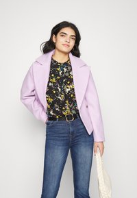 Casaco em lã lilás com lapelas largas, combinado com uma blusa preta de flores e jeans azuis. A modelo segura uma bolsa trançada branca.