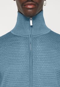 Maglione a collo alto con zip in morbido blu, caratterizzato da un motivo a rilievo, collo alto e cerniera frontale con tirante metallico.