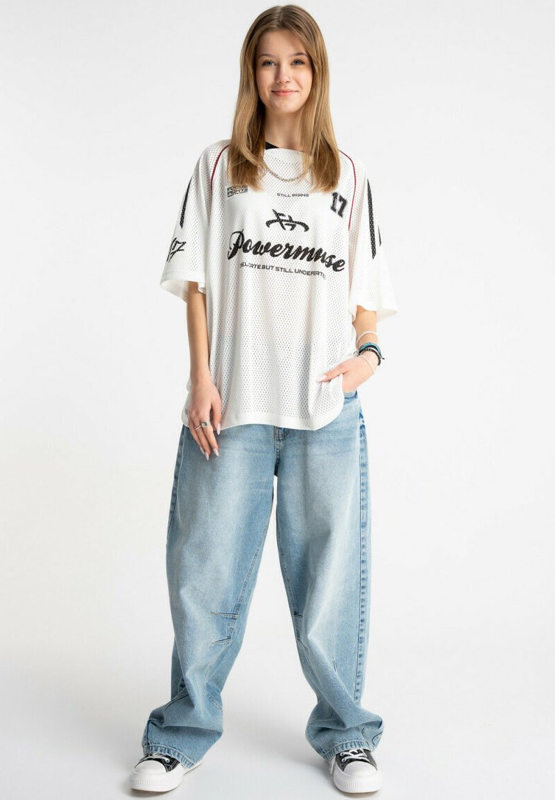 Młoda kobieta stoi uśmiechnięta, ubrana w oversize'ową białą sportową koszulkę, luźne jasnoniebieskie dżinsy i czarno-białe trampki na jednolitym tle.