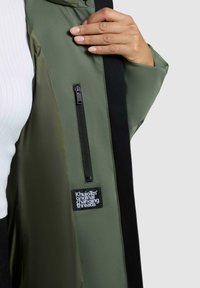 Giacca verde oliva con una tasca con zip e un'etichetta nera con testo bianco. Tessuto morbido e interno foderato, con un design aderente.