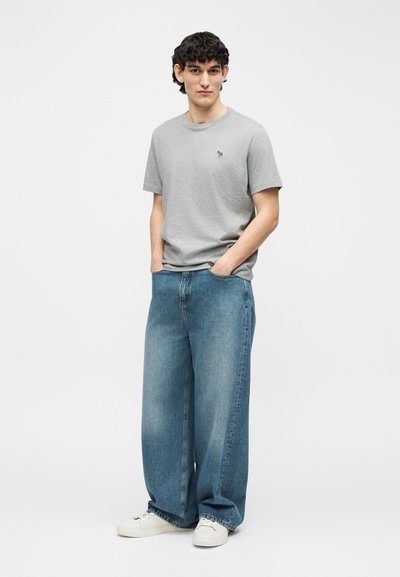 Νεαρός με κατσαρά μαλλιά στέκεται με τα χέρια στις τσέπες, φορώντας γκρι μπλουζάκι, φαρδιά μπλε τζιν και λευκά αθλητικά παπούτσια μπροστά σε λευκό φόντο.