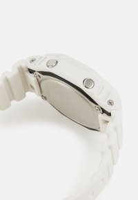 White silicone watch with a metallic silver back, featuring two side buttons and a textured band. The design is angular and thick.