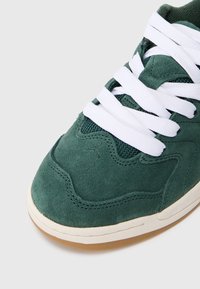 Tenis verde din suede cu sireturi albe, având un design texturat, accente din plasă și o talpă din cauciuc cu un exterior din gumă.
