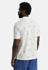 Hvid polo-shirt med krave, prydet med små sejlbådsmønstre i rød og blå. Korte ærmer, slidser i siden og tekstureret stof.