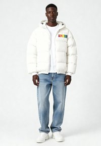 Doudoune blanche à capuche, avec le mot "AMOUR" écrit en lettres colorées sur la poitrine. Portée par-dessus un t-shirt blanc et un jean denim bleu.