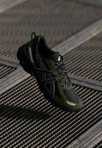Chaussure de sport avec une tige synthétique noire et vert olive, des panneaux en maille texturée, des lacets noirs et un logo d'accent blanc sur le côté.