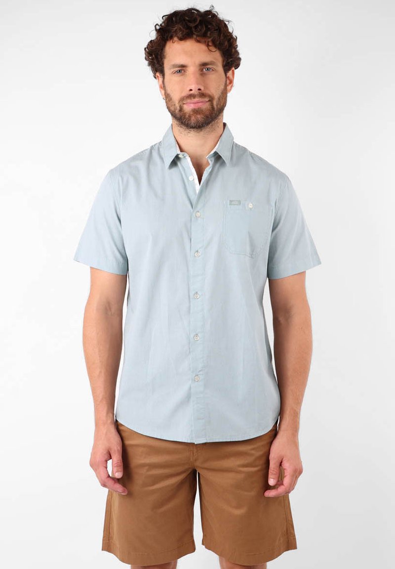 Chemise courte à manches bleues claires avec un col boutonné, poche poitrine simple et texture légère, portée avec un short marron.
