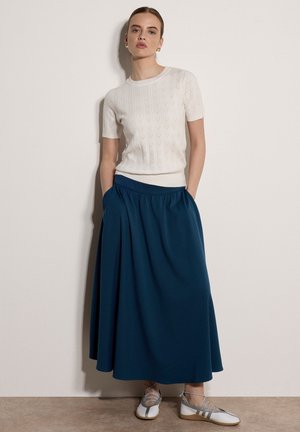 Femme debout contre un mur blanc, portant un top en maille blanc à manches courtes, une longue jupe bleu foncé avec des poches, et des chaussures plates blanches avec des lanières grises.