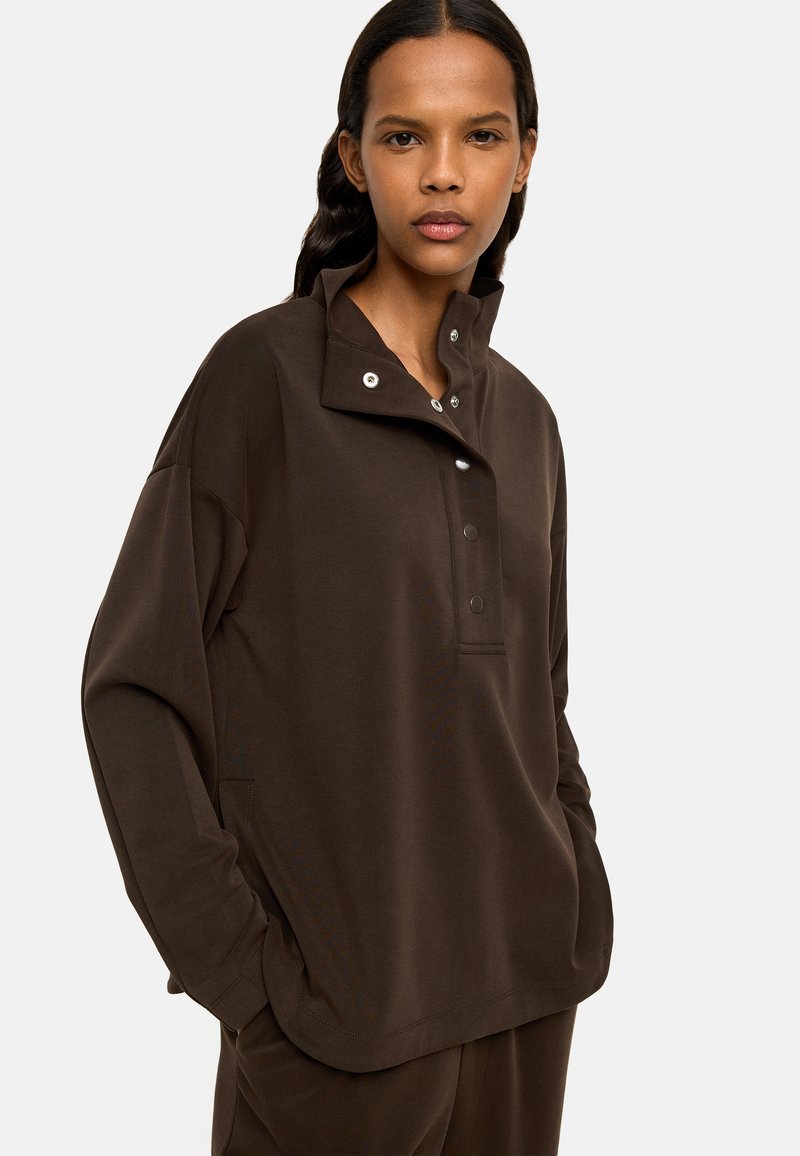 Brązowy sweter z miękkiego materiału, z półkołnierzem, zatrzaskami i bocznymi kieszeniami. Luźny krój z opuszczonymi ramionami i długimi rękawami.