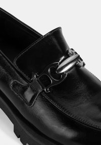 Zwarte leren loafer met een glanzende afwerking, een kettingaccent aan de voorkant en een gestructureerd oppervlak. Chunky zwarte buitenzool.